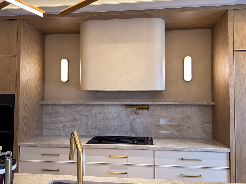 microcement-range-hood-stone-effect-etobicoke-2 A full shot of a kitchen featuring a range hood and surrounding wall finished in a soft beige microcement. The seamless application eliminates crevices, making it highly hygienic and easy to clean. This finish is sealed for stain-resistance, making it an ideal choice for high-traffic food preparation areas in luxury custom homes.
