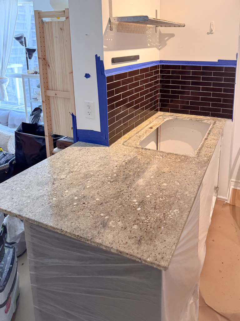 A worn kitchen with existing countertops and a tiled backsplash, showing visible grout lines and signs of age before renovation.