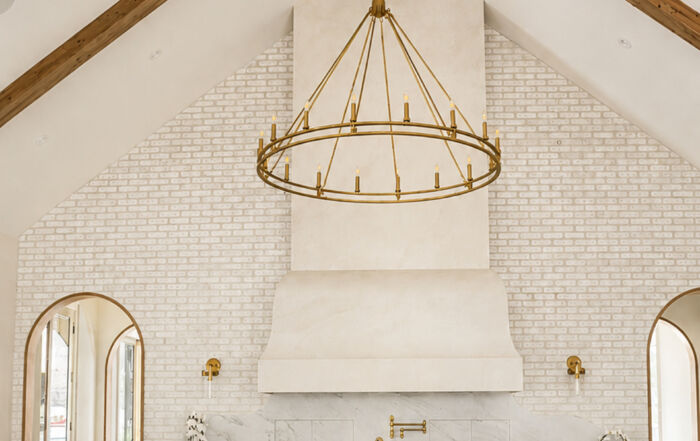 Tall pale plaster range hood against white brick, beneath a vaulted ceiling with exposed timber beams and a gold-toned pendant.